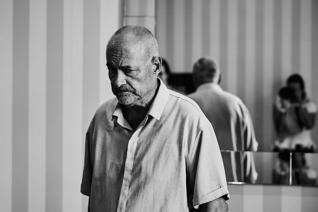 Un homme âgé en chemise claire, tête baissée, devant un mur rayé et un miroir où se reflète une personne qui photographie, en noir et blanc.