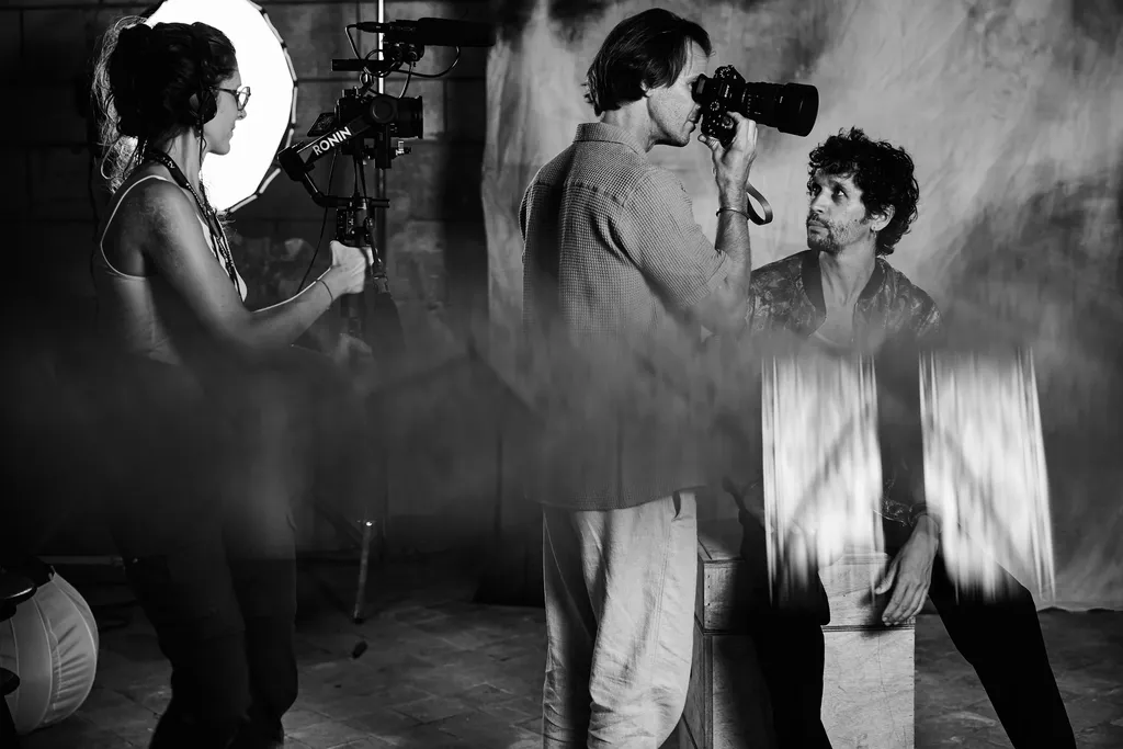 Une cadreuse près d’un projecteur annulaire, un homme photographie un autre homme de près, en noir et blanc.