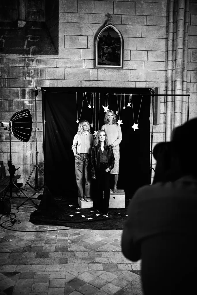 Trois femmes posent sur des caisses devant un fond noir orné d’étoiles suspendues, une boîte à lumière à gauche, dans une salle en pierre, en noir et blanc.