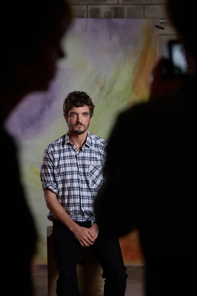 Un homme en chemise à carreaux assis devant une toile peinte, cadré entre deux silhouettes sombres, en couleur.