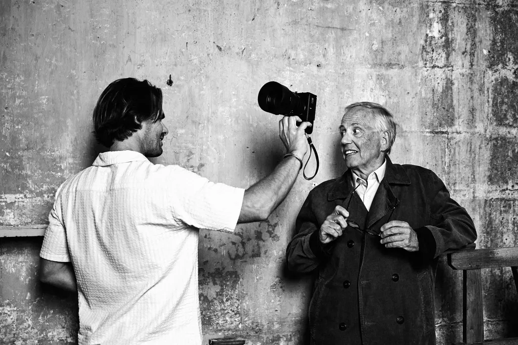 Un homme tend un appareil photo vers un homme âgé en manteau sombre contre un mur usé, en noir et blanc.