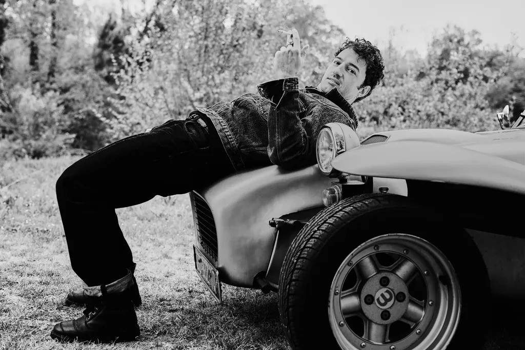 Un jeune homme allongé sur le capot d’une voiture de sport ancienne dans un jardin, en noir et blanc.