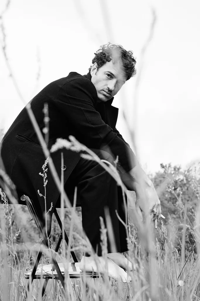 Un homme en manteau noir accroupi sur une chaise pliante, pieds nus dans les hautes herbes, en noir et blanc.