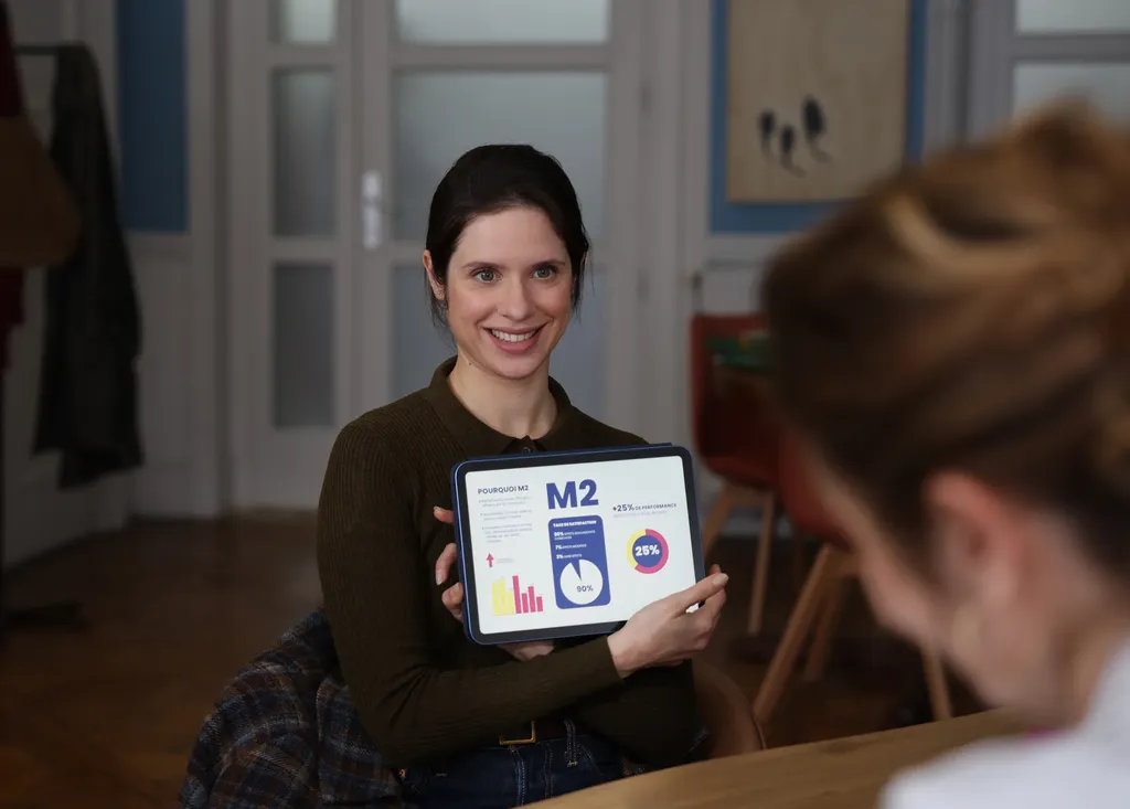 Une femme souriante montre une tablette, vue par-dessus l’épaule d’une autre personne, dans un appartement clair, en couleur.