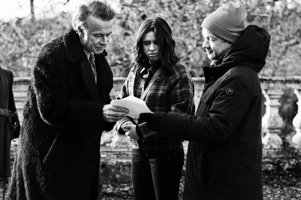 Trois personnes en extérieur lisent une feuille ensemble : un homme en manteau de fourrure, une jeune femme et un homme en bonnet, en noir et blanc.