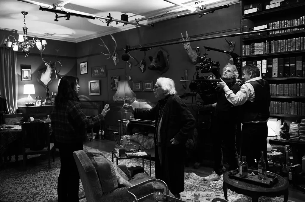 Une équipe tourne dans une bibliothèque ornée de trophées de chasse, caméra sur bras et perche au-dessus des comédiens, en noir et blanc.