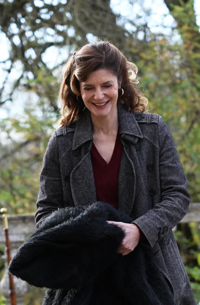 Une femme souriante en manteau gris porte un manteau de fourrure noire sur le bras, arbres d’automne derrière elle, en couleur.
