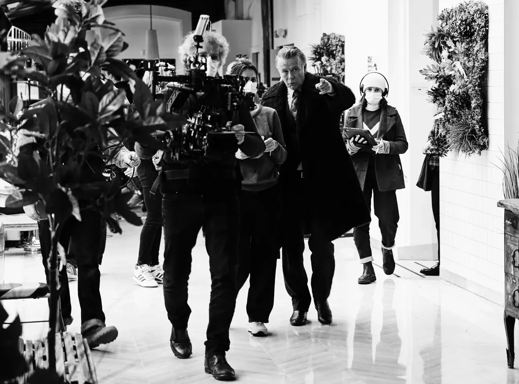 Une équipe avance dans un hall blanc carrelé, un cadreur à l’épaule, un homme qui montre la direction, une femme au casque et à la tablette, en noir et blanc.