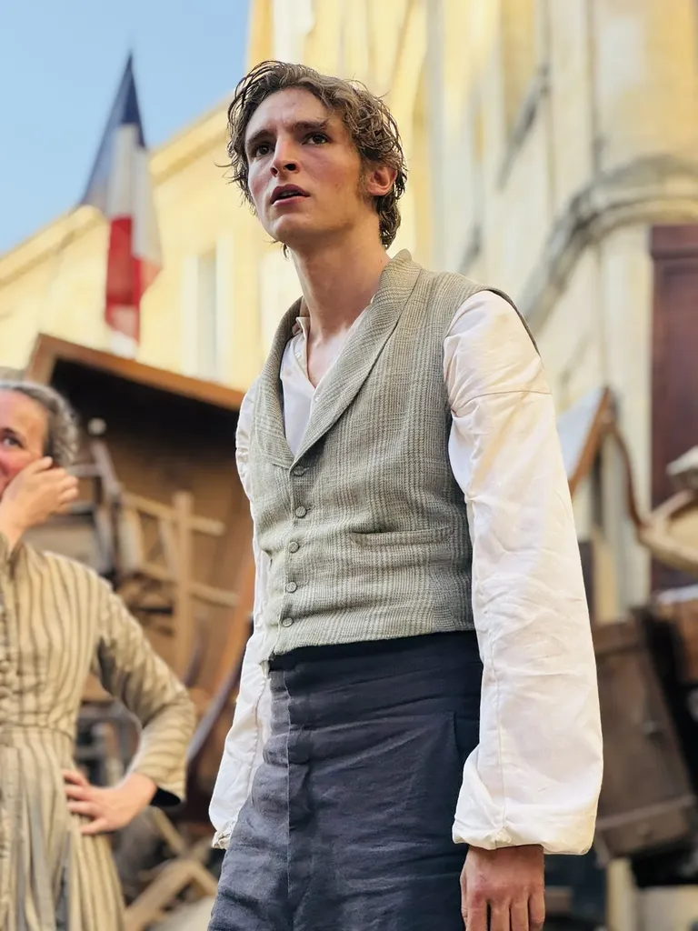 Un jeune homme en costume d’époque, gilet et chemise blanche, dans une rue avec un drapeau tricolore et des débris de bois, en couleur.