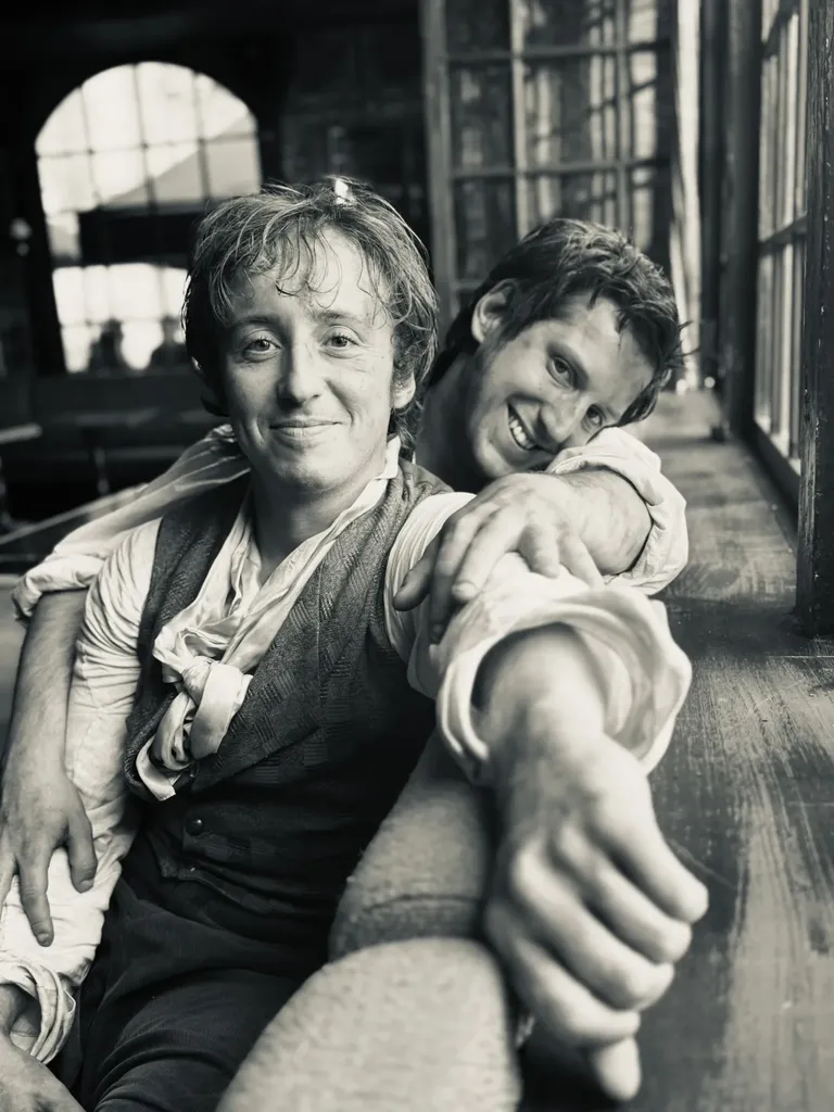 Deux jeunes hommes en costume d’époque, souriants, l’un appuyé sur l’autre près d’une fenêtre, en noir et blanc.