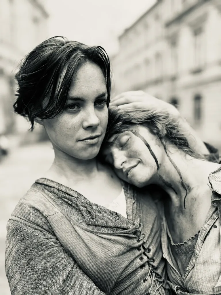 Deux femmes en costume d’époque, l’une pose la tête sur l’épaule de l’autre, visage maquillé de poussière, en noir et blanc.