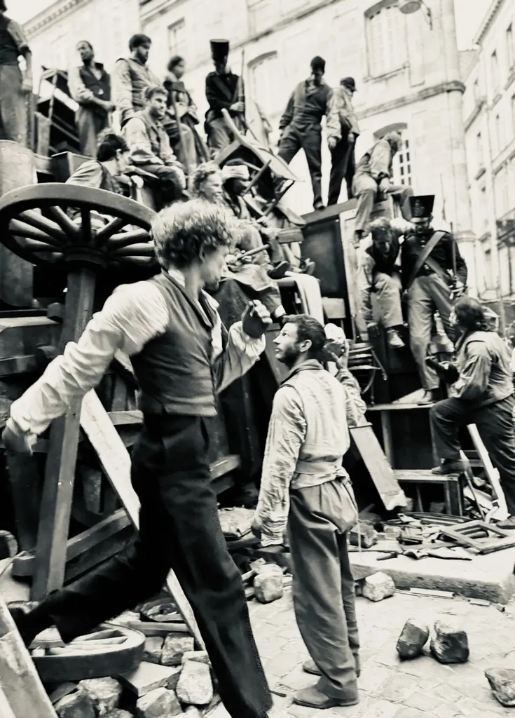 Des figurants en costume d’époque escaladent une barricade de meubles et de roues de charrette dans une rue, en noir et blanc.