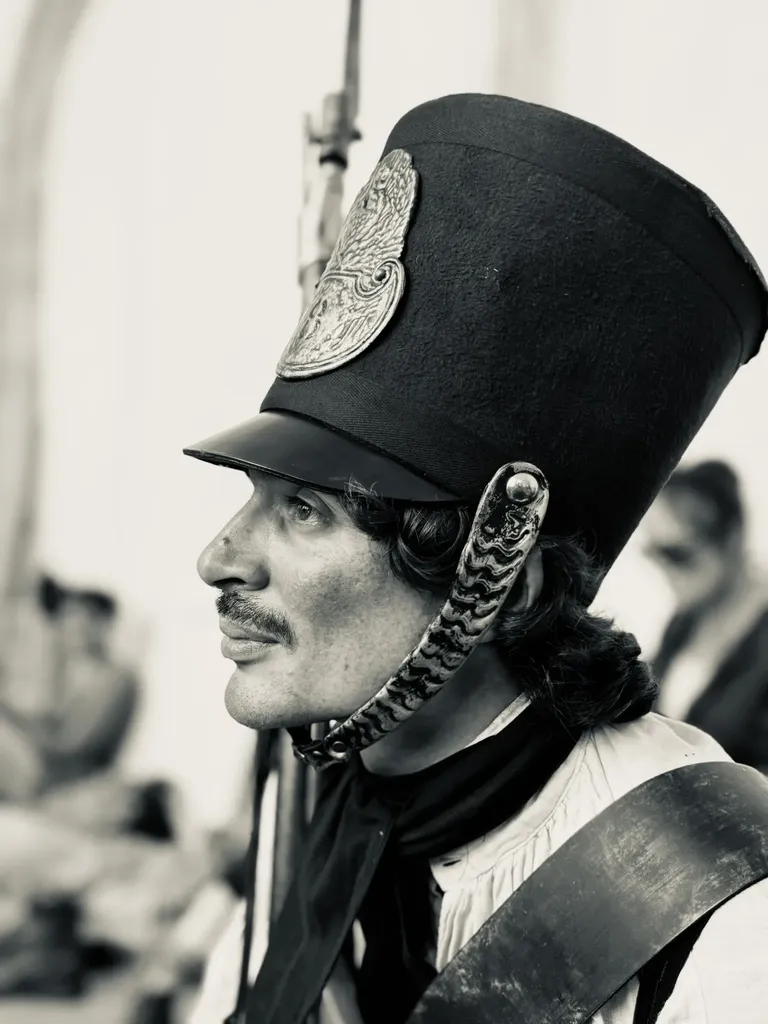 Un soldat de profil coiffé d’un haut shako à plaque de métal et jugulaire, en noir et blanc.