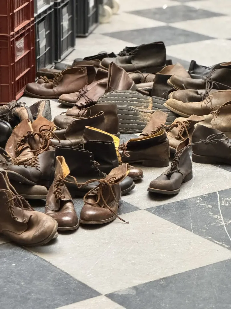 Un tas de chaussures de cuir usées posées sur un sol en damier noir et blanc, près de caisses rouges, en couleur.