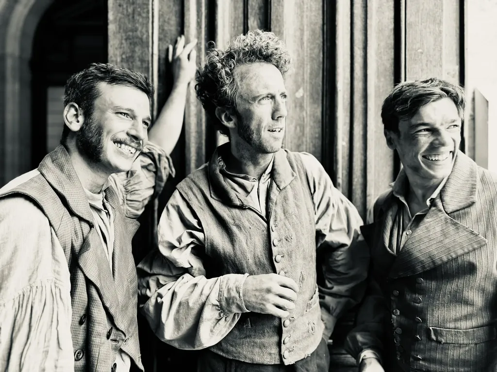 Trois hommes en costume d’époque rient devant une porte en bois, en noir et blanc.