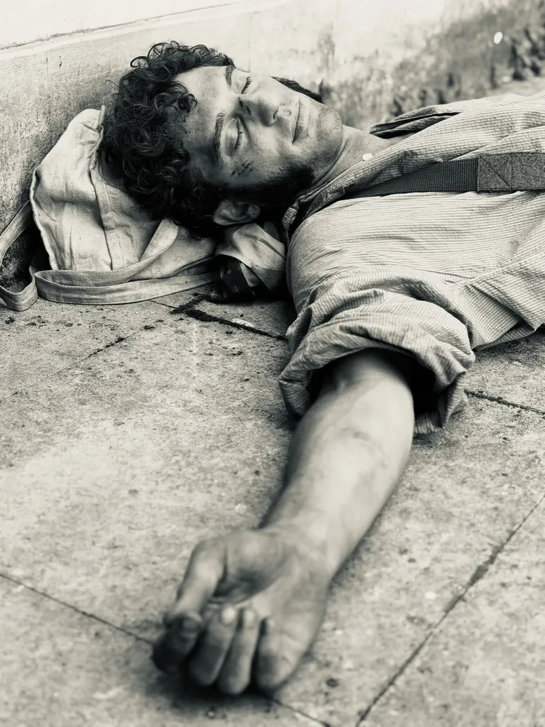 Un jeune homme en chemise d’époque allongé sur les pavés, yeux fermés, le bras tendu, en noir et blanc.