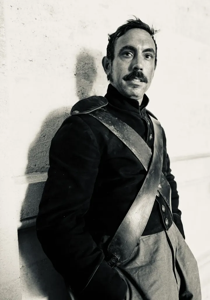 Un homme moustachu en uniforme d’époque à baudrier, adossé à un mur clair, en noir et blanc.