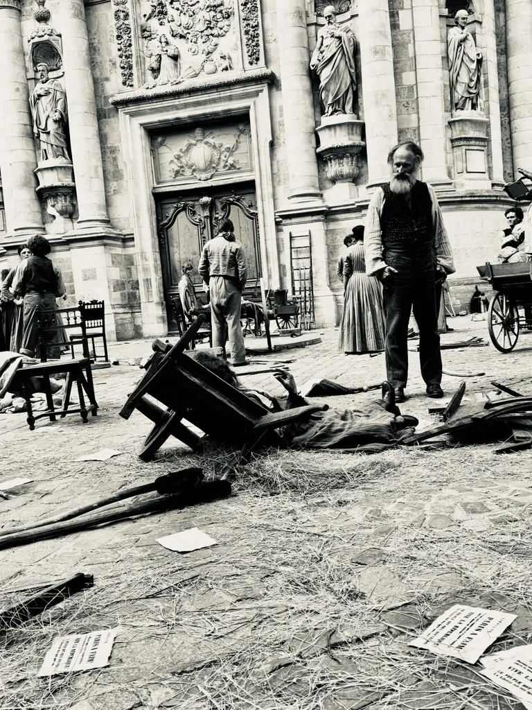 Une place devant une façade d’église sculptée, chaises renversées, paille et papiers au sol, figurants en costume, en noir et blanc.