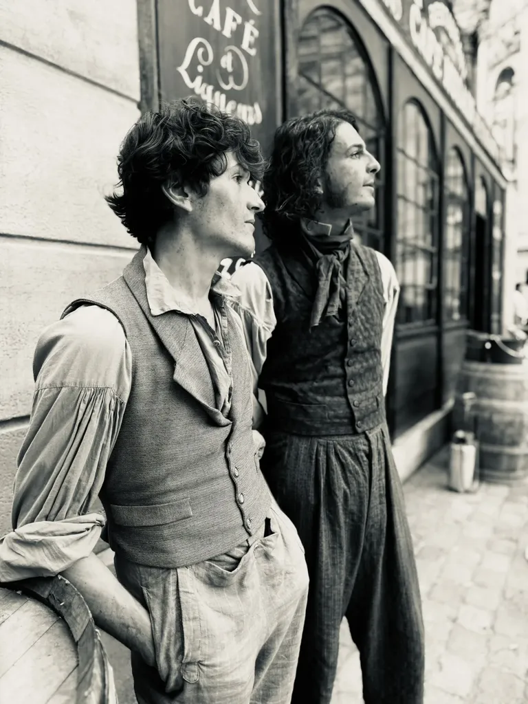 Deux jeunes hommes en gilet devant une devanture de café, le regard tourné vers la droite, en noir et blanc.