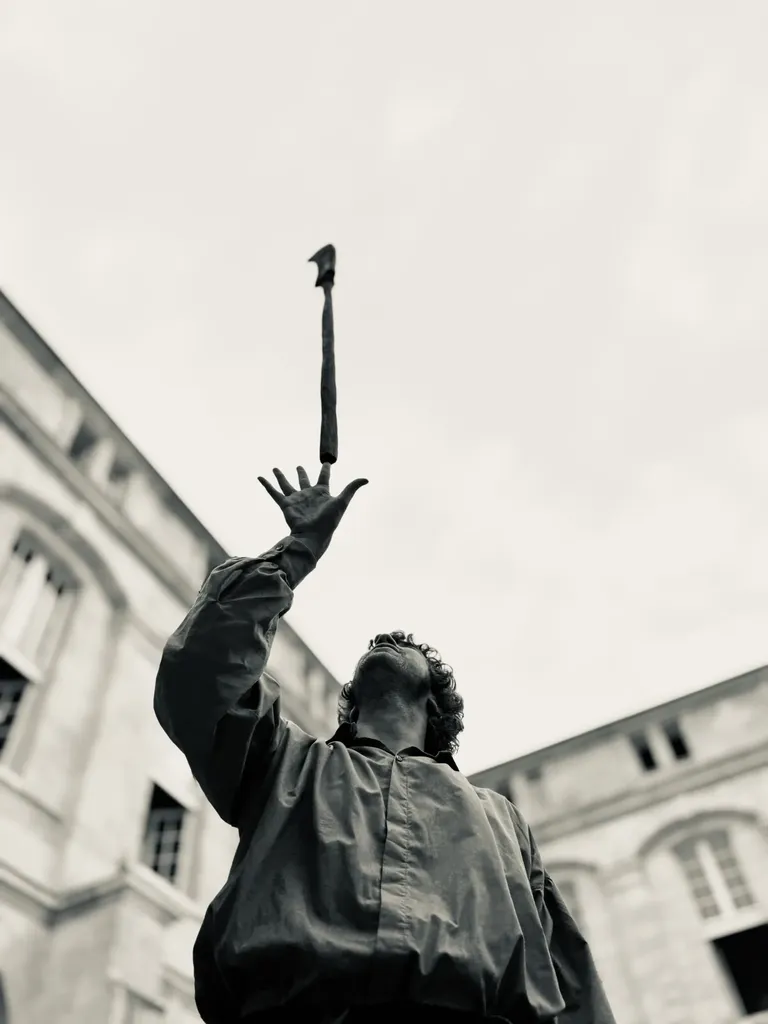 En contre-plongée, un homme en costume tend la main vers un bâton lancé en l’air entre deux façades, en noir et blanc.