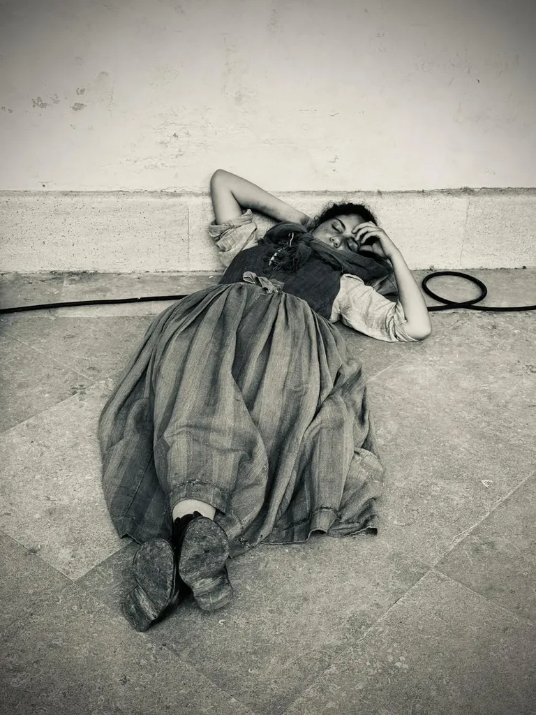 Une femme en longue robe d’époque allongée au pied d’un mur sur un sol de pierre, la main sur le front, un câble à côté d’elle, en noir et blanc.