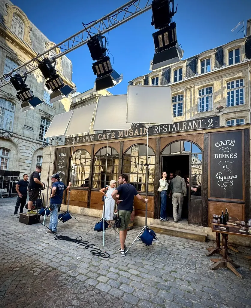 Un décor de café sur une place pavée, une rampe de projecteurs et des cadres de diffusion au-dessus de l’équipe, ciel bleu, en couleur.