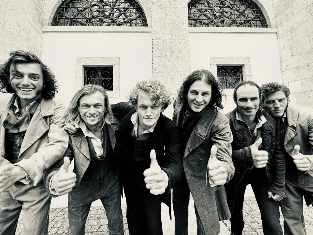 Six hommes en costume d’époque se penchent vers l’objectif, pouces levés, devant un bâtiment de pierre, en noir et blanc.