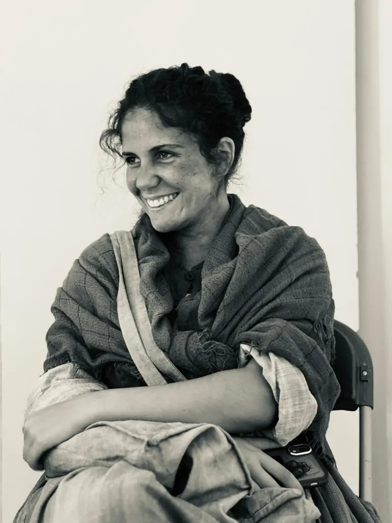 Une femme souriante enveloppée dans un châle, assise sur une chaise pliante, bras croisés, en noir et blanc.