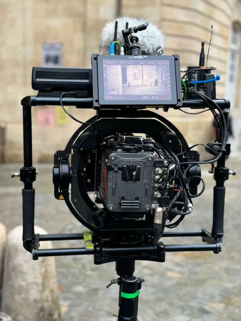 Une caméra de cinéma montée sur un stabilisateur à anneau, écran de contrôle allumé, sur une place pavée, en couleur.