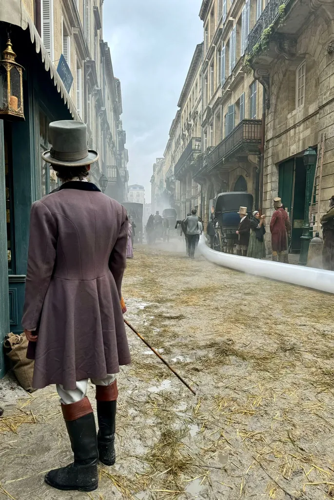 De dos, un homme en redingote prune, haut-de-forme et bottes, avance dans une rue couverte de paille et de fumée, en couleur.