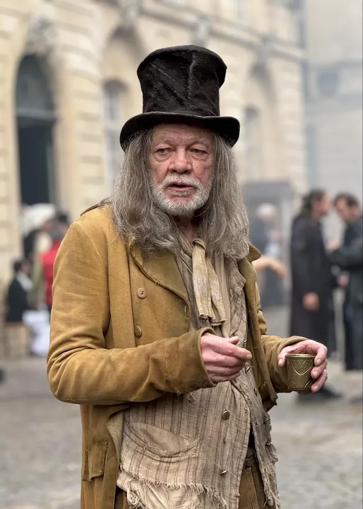 Un homme âgé aux longs cheveux gris et à la barbe blanche, haut-de-forme et manteau ocre usé, tient un gobelet, en couleur.