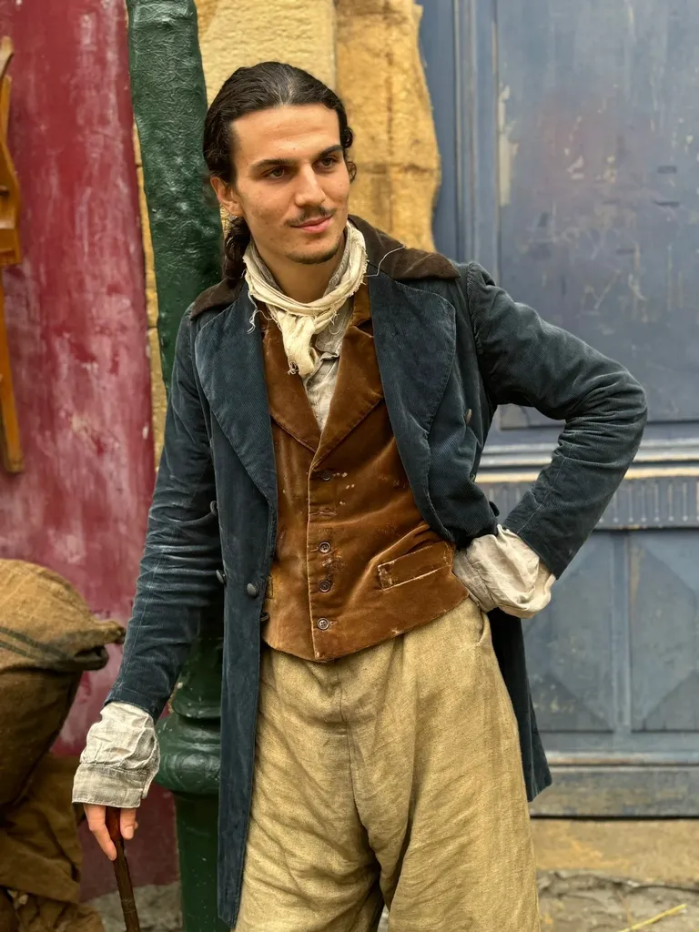 Un jeune homme en redingote de velours et gilet brun, la main sur la hanche, devant un mur rouge et bleu, en couleur.