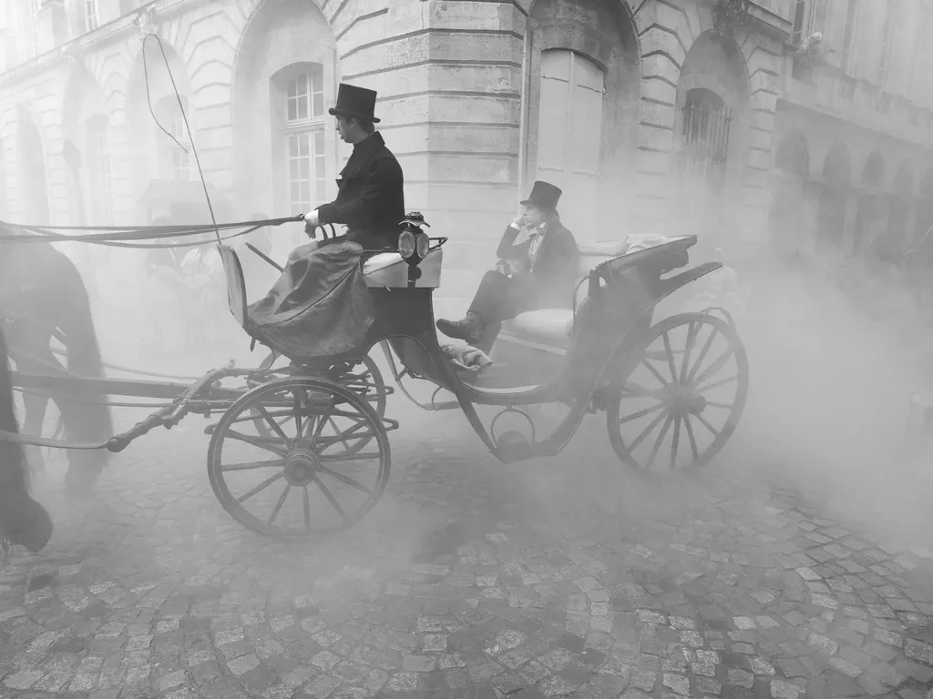 Une calèche avec cocher et passager en haut-de-forme traverse une rue pavée noyée de fumée, en noir et blanc.