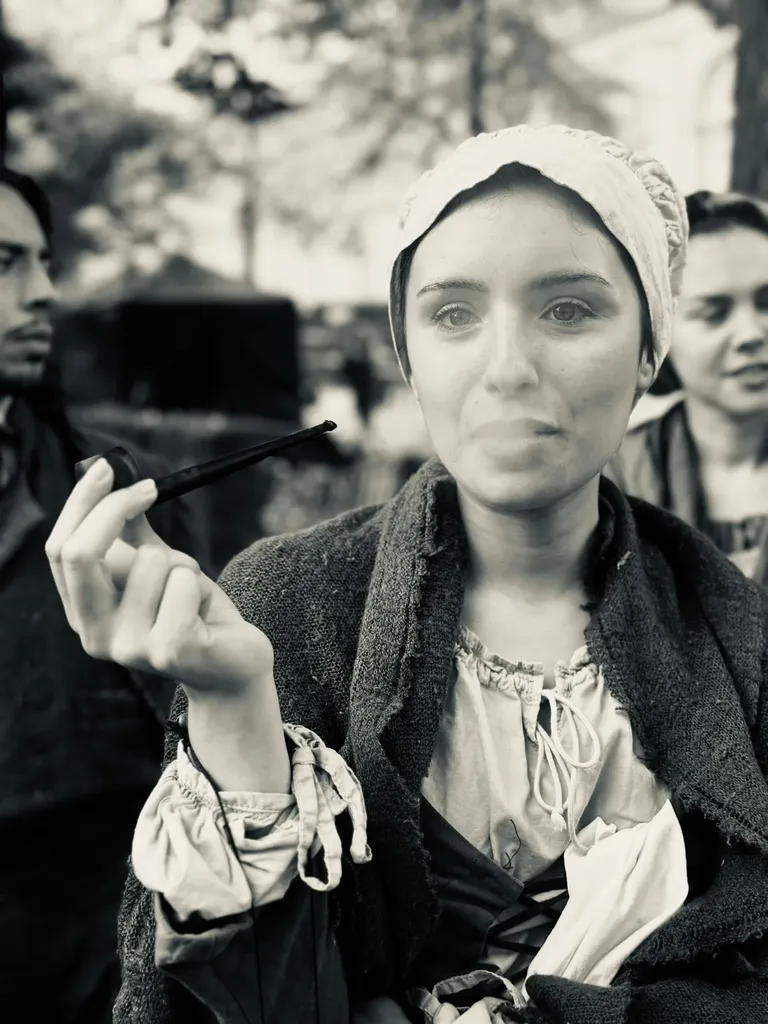 Une jeune femme en bonnet et châle tient une longue pipe et regarde l’objectif, en noir et blanc.