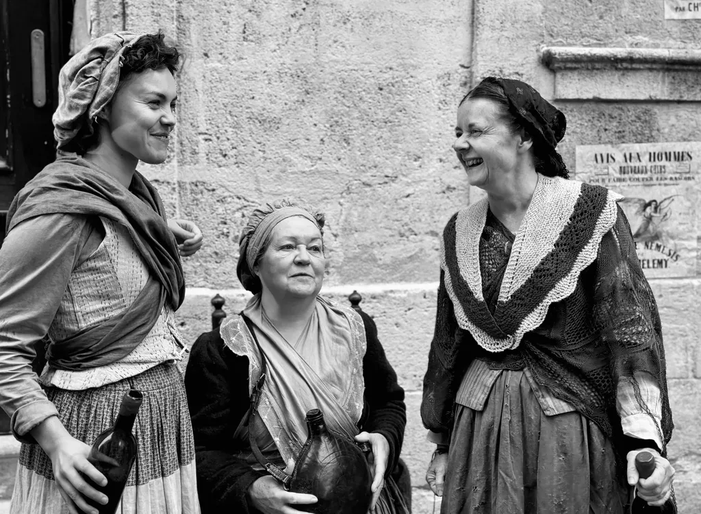 Trois femmes en robes d’époque et foulards rient devant un mur de pierre, en noir et blanc.