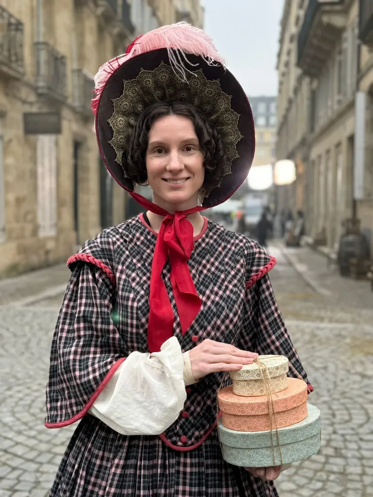 Une femme souriante en robe à carreaux et capote à plume, ruban rouge au cou, tient une pile de boîtes à chapeaux, en couleur.
