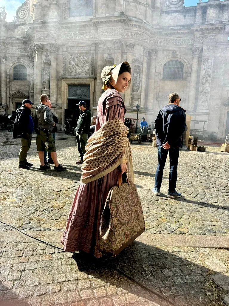 Une femme en robe d’époque et châle se retourne devant une façade d’église sculptée, l’équipe autour d’elle, en couleur.
