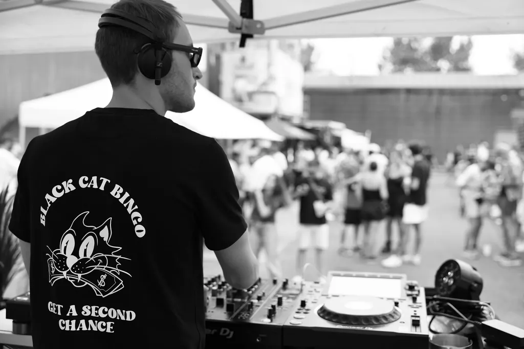De dos, un DJ au casque derrière ses platines sous une tente, face à une foule en plein air, en noir et blanc.