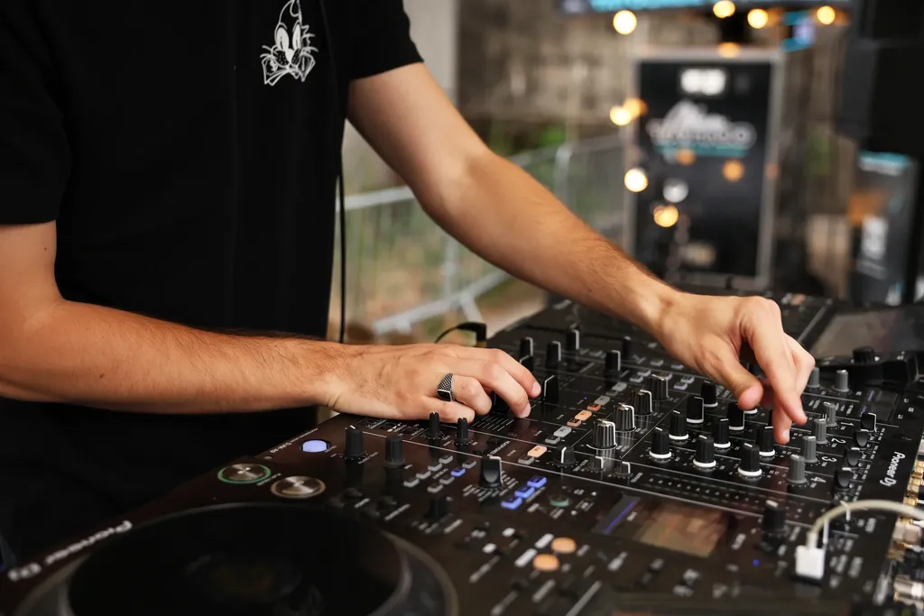 Les mains d’un DJ sur une table de mixage, guirlandes lumineuses floues au fond, en couleur.