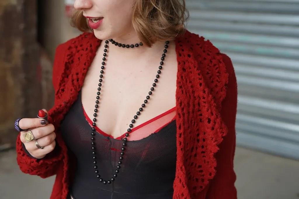 Gros plan sur une femme en gilet rouge au crochet, haut noir, colliers de perles et bagues, lèvres rouges, en couleur.
