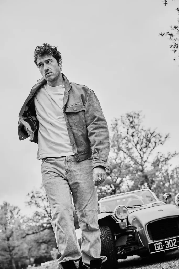 Un homme en veste et jean marche vers l’objectif devant une voiture de sport ancienne, en noir et blanc.
