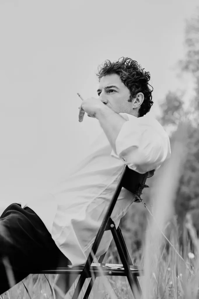 Un homme en chemise blanche assis sur une chaise pliante dans l’herbe, le bras tendu, en noir et blanc.