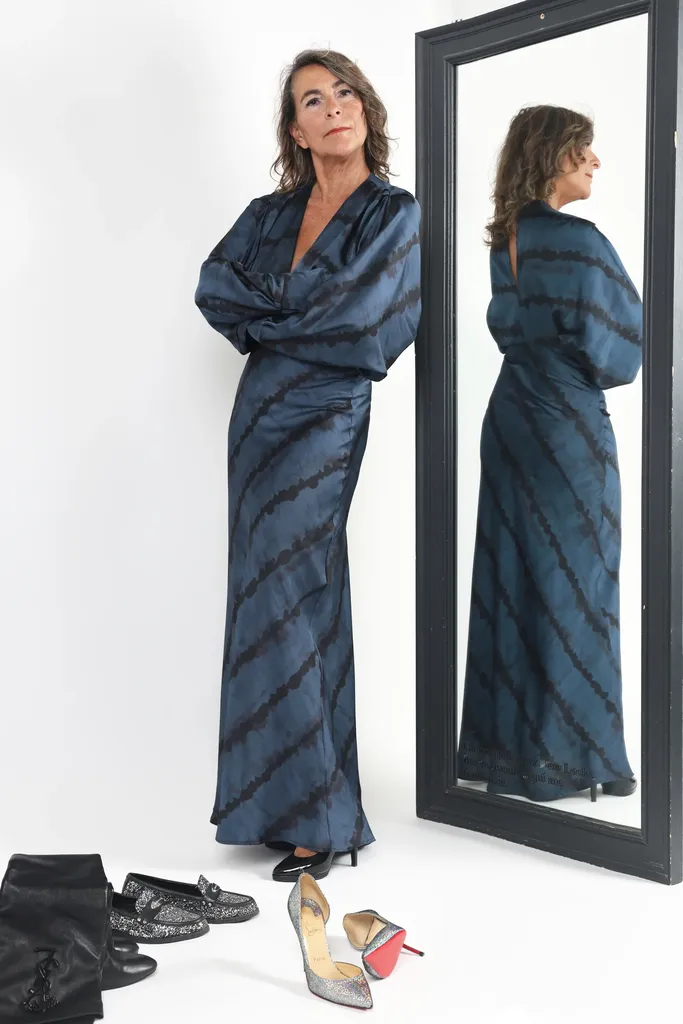 Une femme en robe longue rayée debout près d’un grand miroir où elle se reflète, chaussures posées au sol, studio blanc.