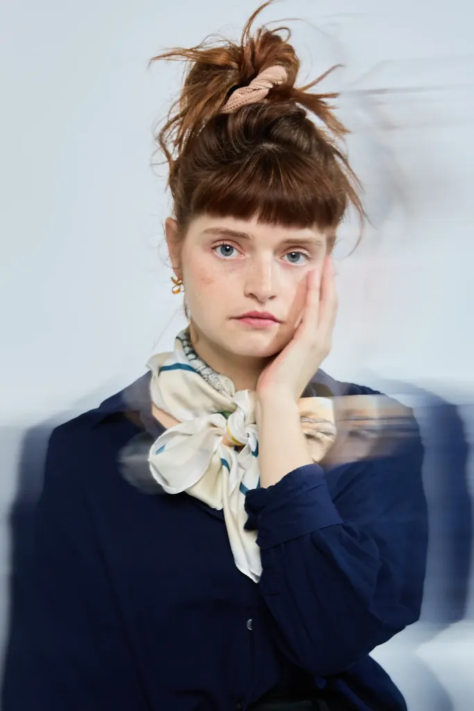 Portrait d’une femme rousse en pull bleu marine et foulard clair, la main sur la joue, fond gris bleu.