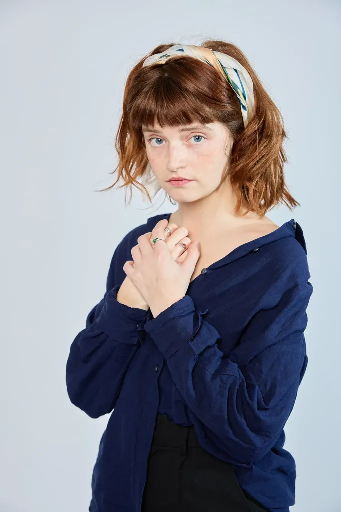 Une femme rousse aux cheveux noués d’un foulard, mains croisées sur la poitrine, fond gris bleu.