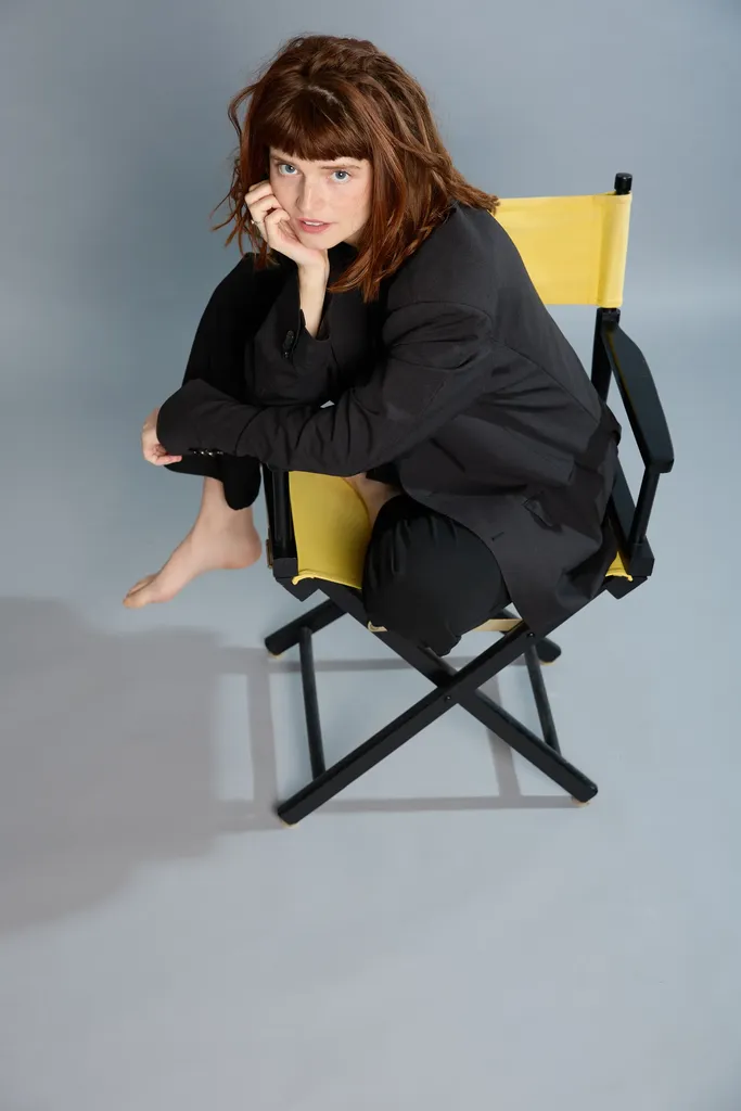 Une femme rousse assise sur une chaise de réalisateur à toile jaune, le menton dans la main, fond gris bleu.