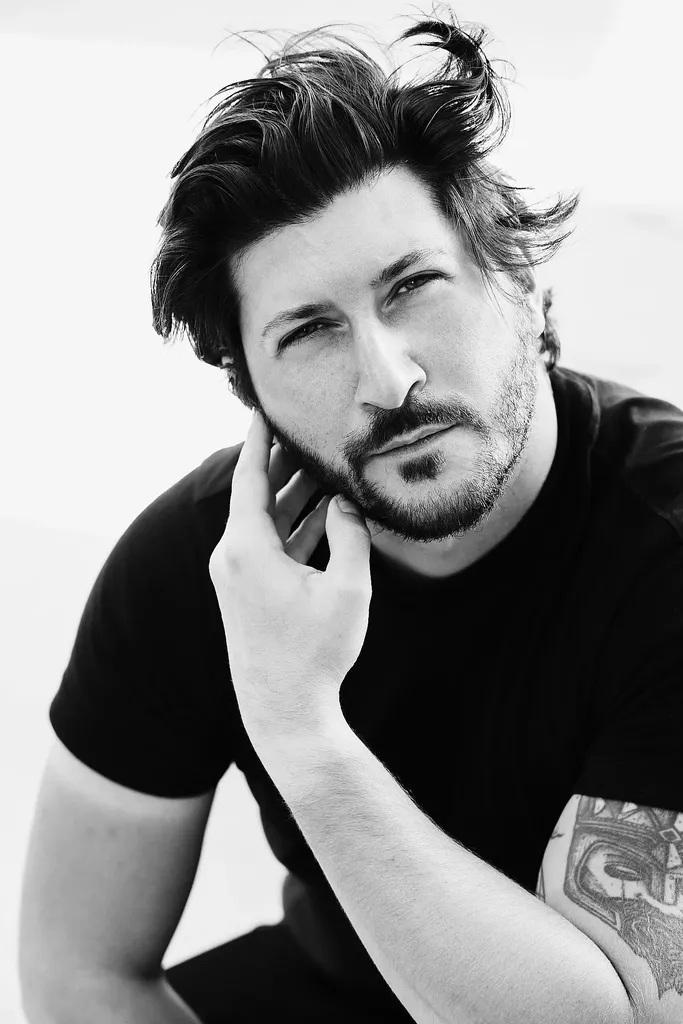 Un homme barbu en t-shirt noir, la main près du visage, portrait serré en noir et blanc.