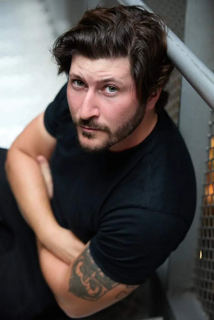 Un homme barbu en t-shirt noir, bras croisés près d’une rambarde, vu en plongée, en couleur.