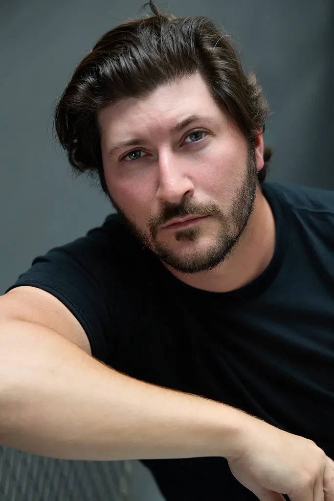 Un homme barbu en t-shirt noir accoudé, regard droit vers l’objectif, fond gris sombre, en couleur.