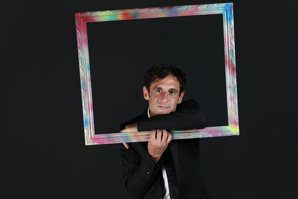 Un homme en costume sombre tient un cadre multicolore devant lui, sur fond noir, cadrage horizontal.